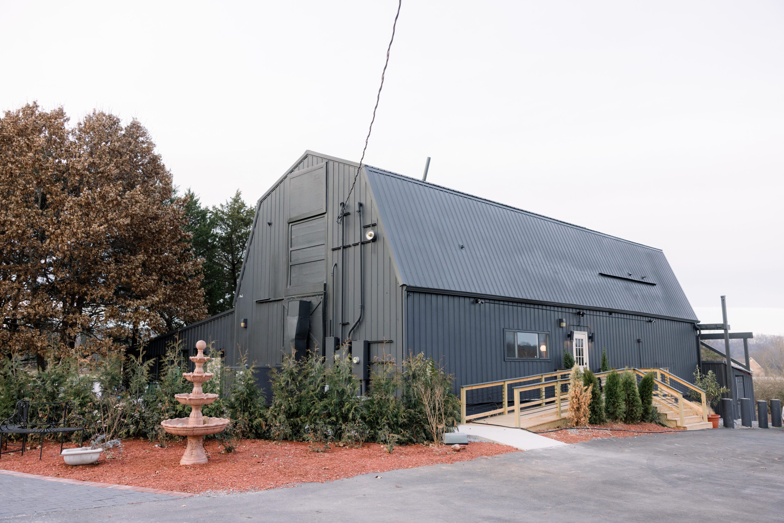 Large gray barn with a ramp and surrounding plants.
