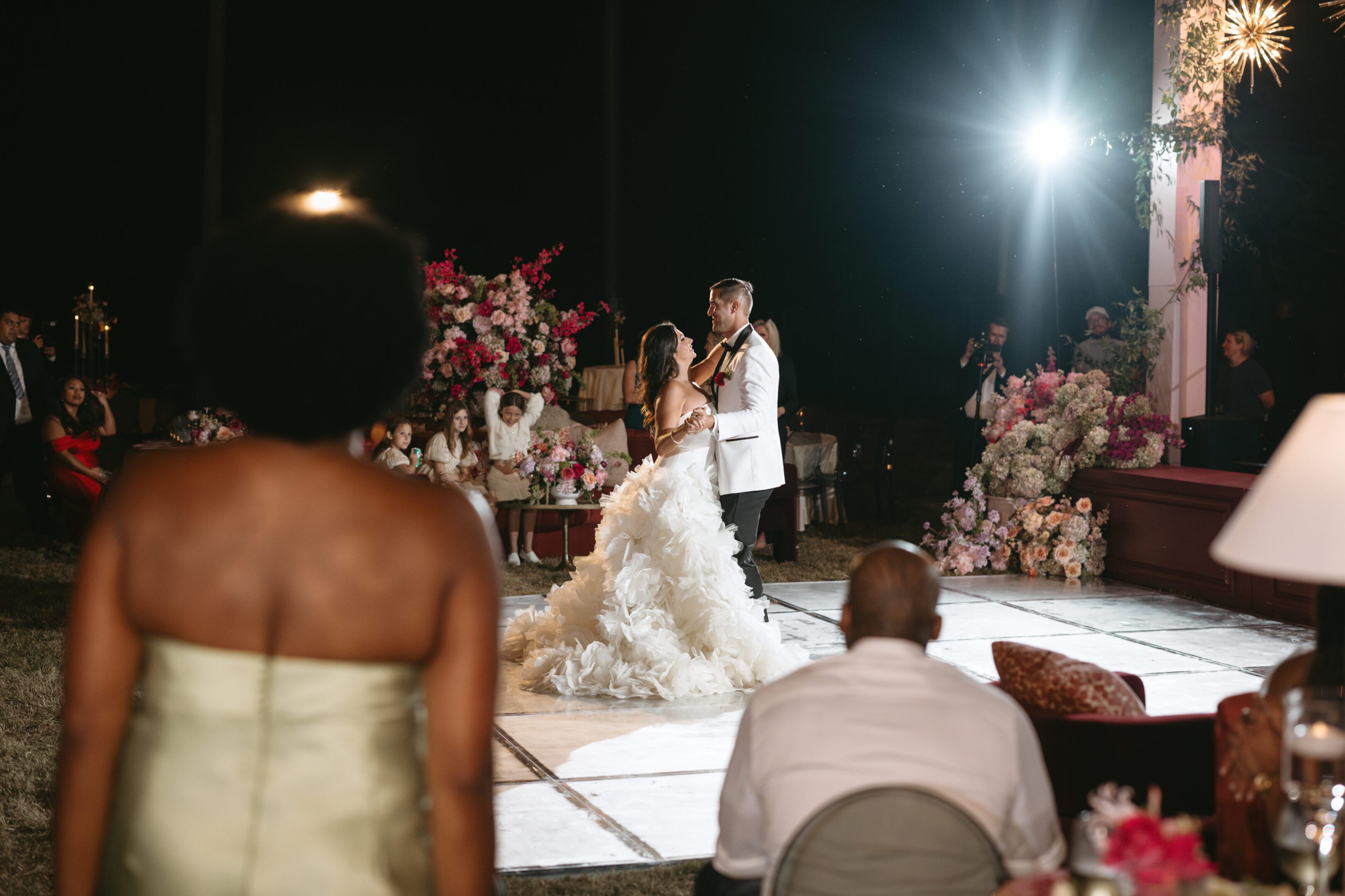 A couple shares a dance surrounded by guests and floral decorations at night.
