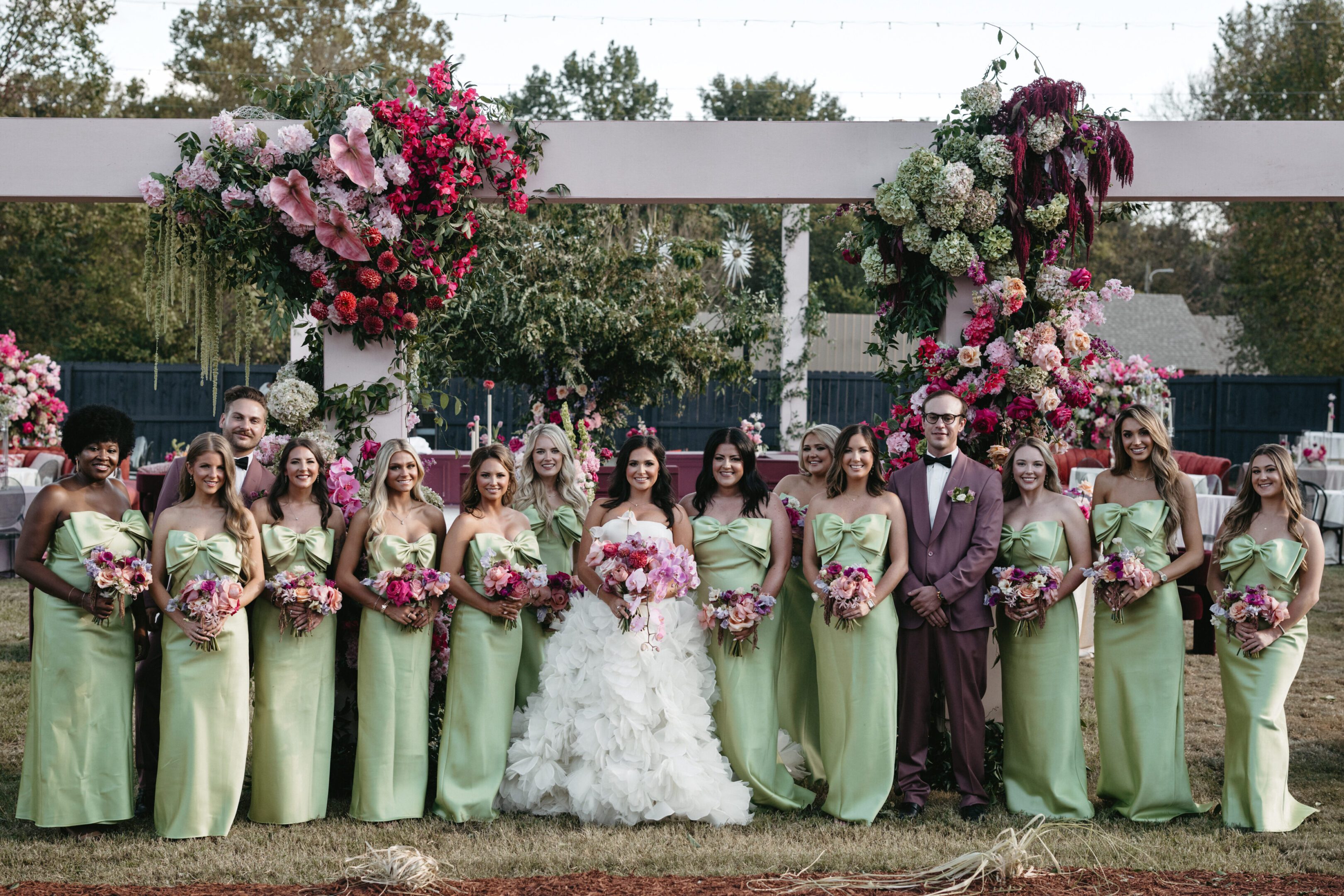 Bride with bridesmaids in green dresses holding pink bouquets outdoors.