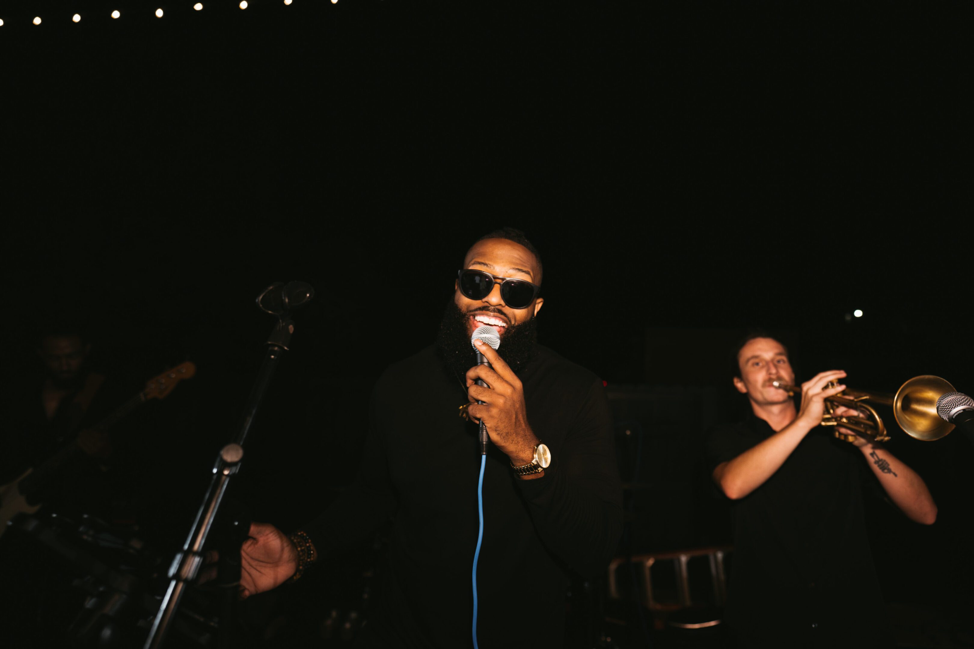 A performer singing into a microphone on stage wearing sunglasses.