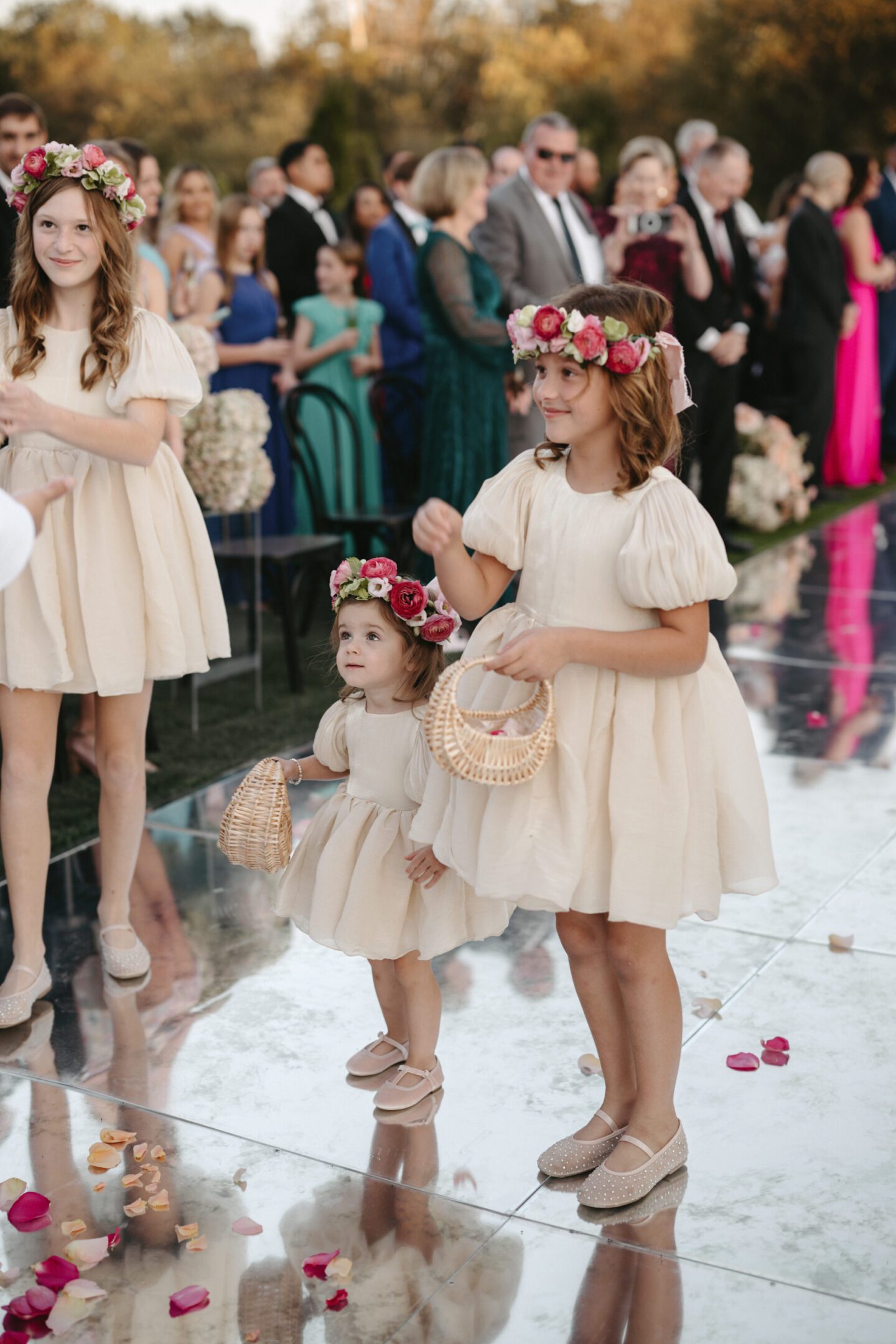 Three young girls in matching cream dresses with flower crowns at an event.
