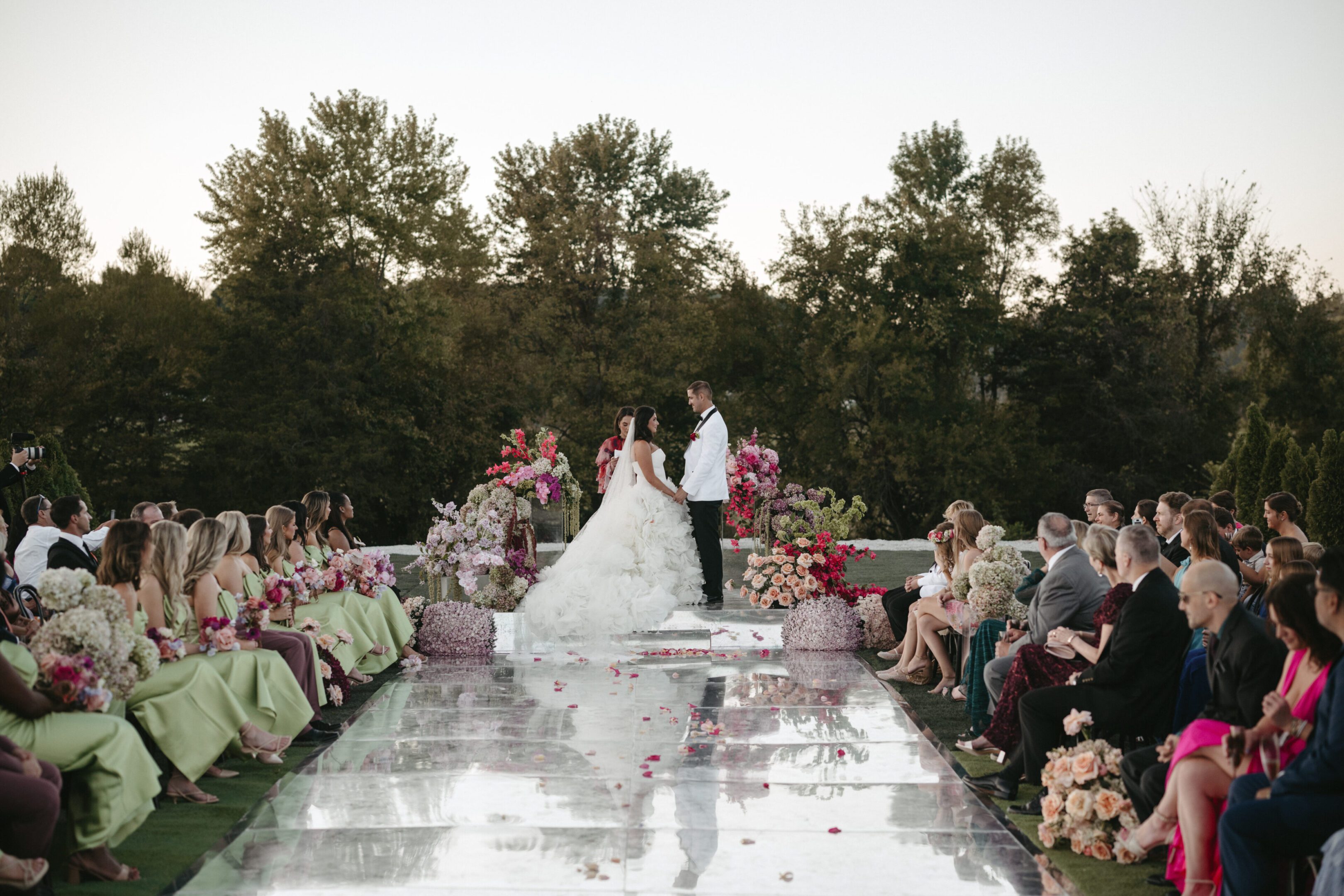 Bride and groom walk down a reflective aisle surrounded by guests and floral arrangements outdoors.