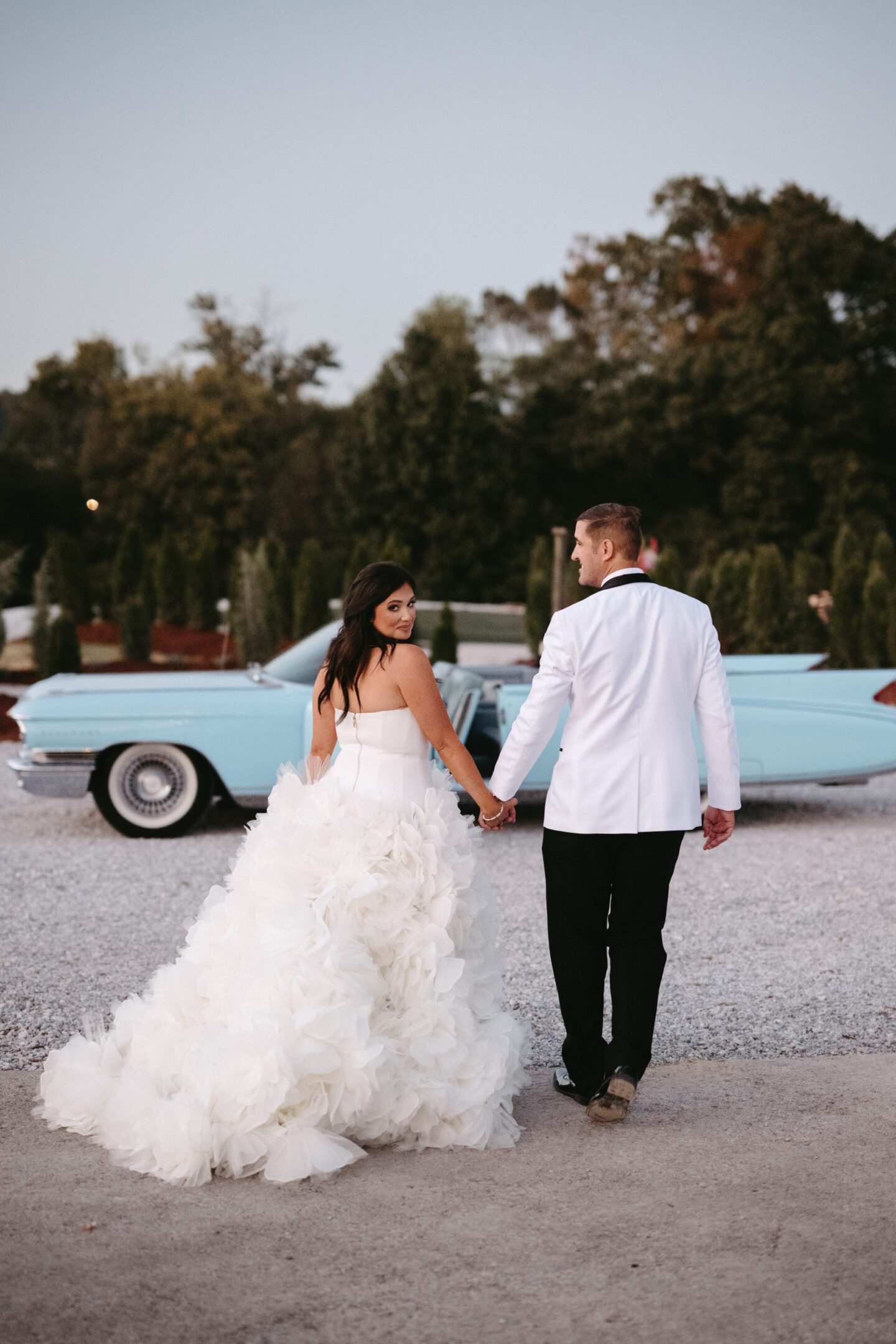 Bride and groom walking hand in hand near a classic blue car.