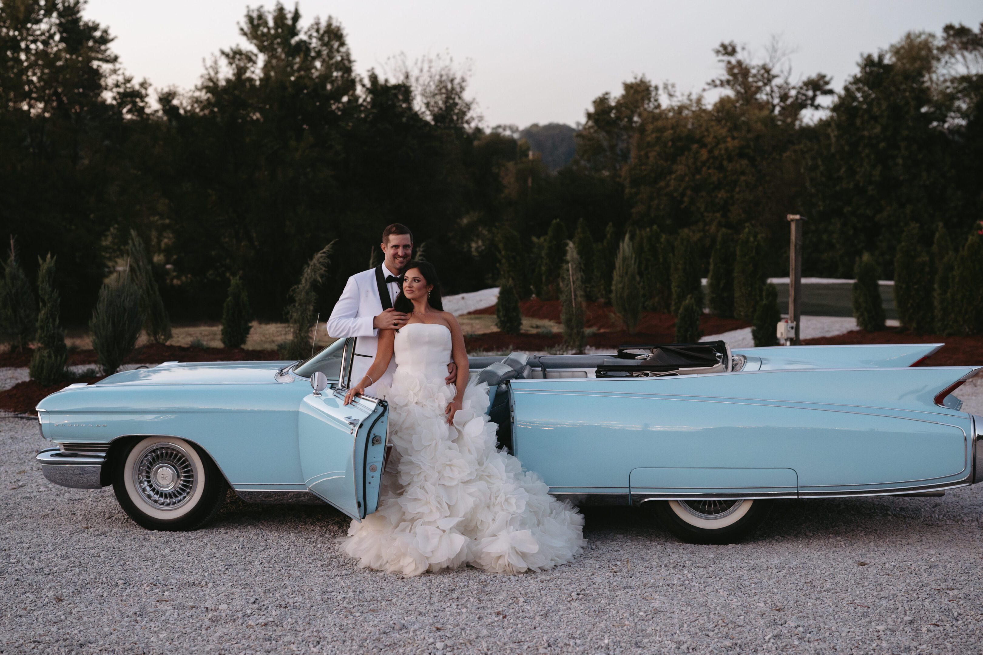 Bride and groom posing by a vintage blue car outdoors.
