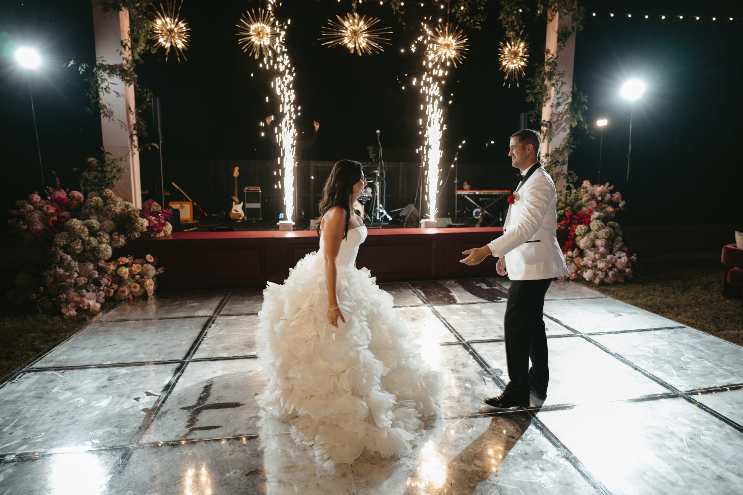 A bride and groom stand facing each other with celebratory sparks around them.